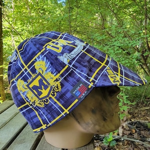 May include: A cycling cap featuring a blue, black, and yellow plaid pattern with University of Michigan Wolverines logos and text. The cap includes a small black tag on the side that reads Made in U.S.A. with a small American flag graphic.
