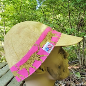 May include: A tan visor with a pink band featuring a leopard print. The visor has a curved brim and is labeled "MADE IN USA" on a small tag.