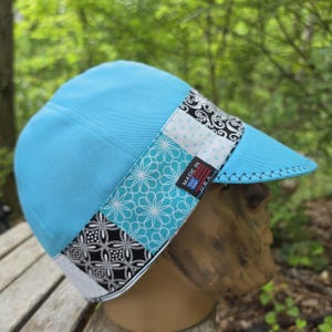 May include: A light blue welding cap with a patterned band featuring white, black, and turquoise floral and geometric designs. The cap has a curved brim and a label that reads "Made in U.S.A."