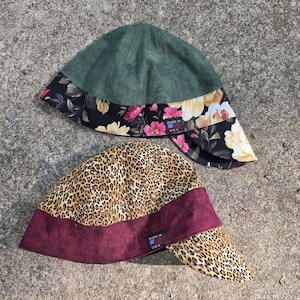May include: Two welder's caps. One is green with a floral print brim featuring pink and yellow flowers on a black background. The other is leopard print with a burgundy brim. Both have a "Made in USA" tag.