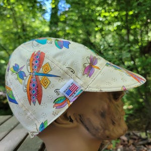 May include: A cream-colored newsboy cap featuring a colorful dragonfly and butterfly pattern. The cap has a small brim and is adorned with a "Made in U.S.A." tag. The dragonfly and butterfly designs are in shades of blue, orange, purple, and green.