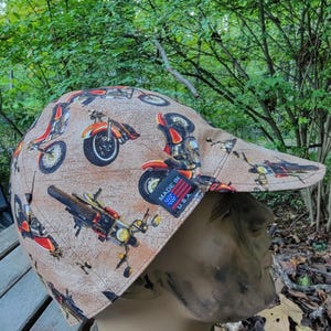 May include: A tan cap featuring a repeating pattern of motorcycles in red, orange, and black. The cap has a curved brim and a tag that reads "MADE IN U.S.A."