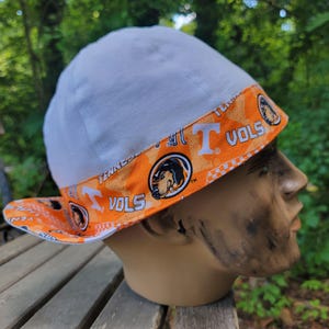 May include: A white and orange hat with the text "VOLS" and a graphic of a lion. The hat is on a mannequin head. The hat has a white top and an orange brim with the team's logo.