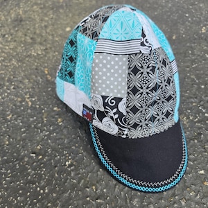 Könnte beinhalten: Eine Schweißerkappe mit Patchwork-Design aus schwarzen, weißen und türkisfarbenen Stoffmustern, darunter florale, gepunktete und geometrische Motive. Die Kappe hat einen einfarbig schwarzen Schirm mit türkisfarbenen Nähten und ein Etikett Made in USA.