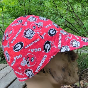 May include: A red baseball cap featuring the University of Georgia Bulldogs logo and the word "GEORGIA" in white. The cap has a curved brim and is made of a patterned fabric.