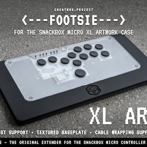 May include: Black and clear rectangular controller with black buttons and the text "FOOTSIE" and "XL ART". Features include wrist support, a textured baseplate, and cable wrapping support. Designed for the Snackbox Micro XL artwork case.
