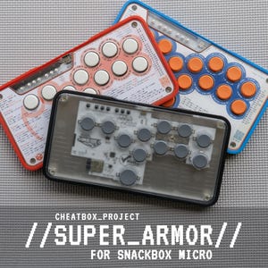 May include: Three rectangular game controllers with button layouts. One has a red frame with white buttons, one has a blue frame with orange buttons, and the front one has a black frame with grey buttons and a transparent top cover showing internal circuitry.
