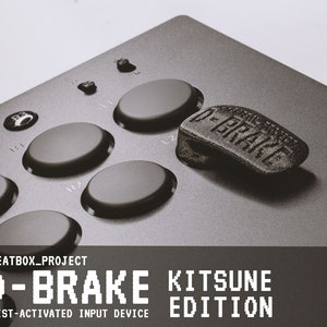 Może przedstawiać: Czarno-szare urządzenie wejściowe aktywowane nadgarstkiem, D-BRAKE Kitsune Edition, z serią okrągłych przycisków i teksturowaną podkładką z logo D-BRAKE. Urządzenie jest przeznaczone do gier i ma tekst "CHEATBOX_PROJECT" na dole.