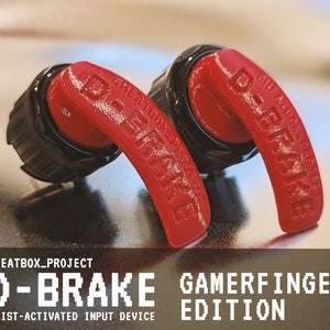 May include: Two red D-Brake Gamerfinger Edition wrist-activated input devices. The devices are made of red plastic and have a black base. The text "CHEATBOX_PROJECT" and "D-BRAKE GAMERFINGER EDITION" is printed on the devices.