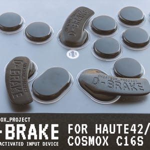 May include: An overhead shot of a gray surface with several black, circular buttons and D-BRAKE components. The text "CHEATBOX_PROJECT" and "D-BRAKE" is visible, along with "FOR HAUTE42/ COSMOX C16S WRIST-ACTIVATED INPUT DEVICE".