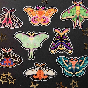 Puede incluir: Una colección de pegatinas de polillas coloridas con diseños intrincados. Las pegatinas presentan varios colores, incluyendo rosa, naranja, verde y azul, con contornos negros. Confeti dorado en forma de estrella está esparcido alrededor de las pegatinas.