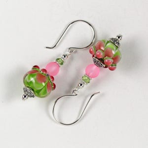 May include: A pair of handcrafted earrings featuring glass beads in shades of pink, green, and red. The earrings have silver-colored ear wires and decorative metal accents. The design includes round pink beads, green and red patterned beads, and small green beads.