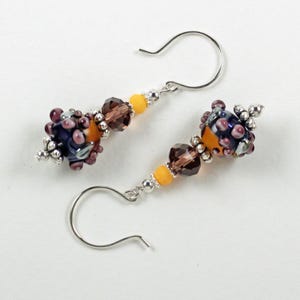 May include: A pair of handcrafted earrings featuring dark blue and orange glass beads with purple accents. The earrings also include brown and yellow beads, and silver-colored ear wires. The design is intricate and detailed, showcasing a unique style.