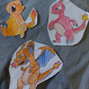 以下が含まれることがあります： 白い紙に手書きされたポケモンのキャラクターが3体。キャラクターはヒトカゲ、リザード、リザードン。ヒトカゲはオレンジ色で炎の尻尾、リザードはオレンジ色で青い炎の尻尾、リザードンは赤色で青い炎の尻尾です。