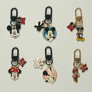 May include: Six different keychains featuring Mickey Mouse and Minnie Mouse. The keychains are made of metal and have a gold finish. Each keychain has a different design, including Mickey Mouse with a red bow tie, Minnie Mouse with a pink bow, and Mickey Mouse with a blue bow tie. The keychains are all attached to black metal rings.