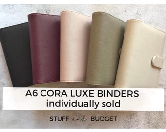 Op de afbeelding: Vijf A6 Cora Luxe binders in verschillende kleuren: zwart, bordeaux, blush, olijfgroen en crème. De binders worden gepresenteerd op een lichtgekleurd oppervlak. Tekst op de afbeelding luidt "A6 CORA LUXE BINDERS individually sold" en "STUFF and BUDGET".