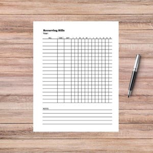May include: A white sheet of paper with a grid for tracking recurring bills, with the title "Recurring Bills" at the top. A black and silver pen rests on the right side of the paper. The background is a light brown wooden surface.