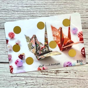 May include: A white gift card with origami cranes and gold circles. The card features the text "stuff and budget" and the value "£50". Decorative paper balls in pink, purple, and red are scattered around the card.
