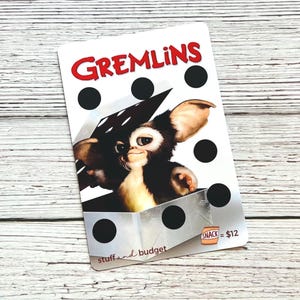 May include: A white card featuring the word "GREMLINS" in red, with a cartoon gremlin character. The card has black dots and the text "stuff and budget". The image also includes the text "SNACK = $12".