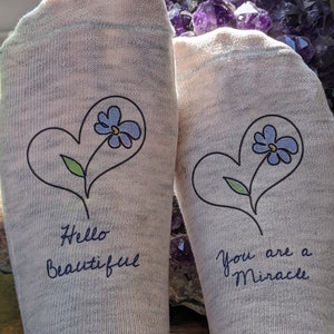 May include: A pair of white socks with blue flower designs and handwritten messages. One sock says "Hello Beautiful" and the other says "You are a Miracle".