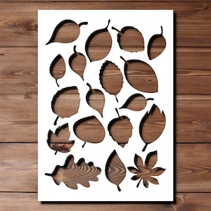 May include: A white stencil sheet featuring a variety of leaf shapes cut out, displaying a wood grain pattern. The leaves are of different sizes and styles, some with detailed features. The background is a wooden surface.