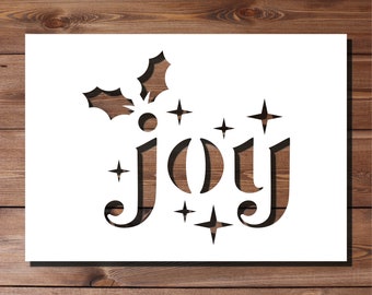 May include: A white stencil with the word "JOY" cut out, featuring a holly sprig and starbursts. The background shows a wooden surface. The letters and design elements are cut out, revealing the wood grain.
