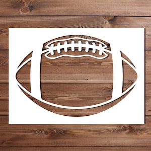 May include: A white football stencil cut-out, featuring the shape of a football with detailed stitching. The stencil is set against a wooden background, creating a contrast between the white design and the brown wood.