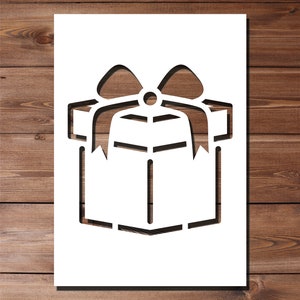 Puede incluir: Una plantilla blanca de una caja de regalo con un lazo en la parte superior. La plantilla está sobre un fondo de madera marrón.