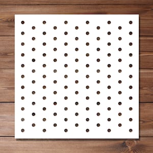 May include: A white stencil featuring a repeating pattern of dark brown polka dots, evenly spaced across the surface. The square stencil is placed on a wooden background with a warm, brown colour.