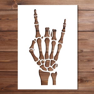 May include: A white stencil featuring a die-cut design of a human skeletal hand making the rock and roll sign. The hand is shown with the index and pinky fingers extended upward while the middle and ring fingers are folded down toward the palm.