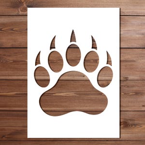 May include: A white rectangular stencil featuring a large bear paw print cutout. The design includes a central pad and five distinct toe pads with sharp claw marks at the top, creating a clear silhouette of a wild animal track for crafting.