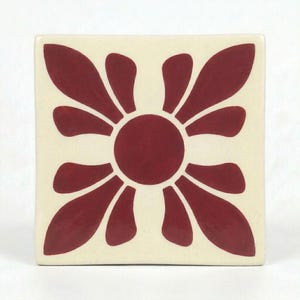 May include: A square ceramic tile featuring a cream-colored background with a central dark red circular shape surrounded by eight symmetrical, petal-like forms that extend toward the corners and edges of the tile in a repeating pattern.