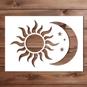 May include: A white stencil design of a sun, crescent moon, and three stars. The sun has curved rays, and the moon is a cutout shape. The design is set against a wooden background, creating a contrast between the white stencil and the wood grain.
