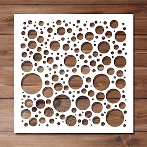 May include: A white stencil with a pattern of varying sized circles. The stencil is square and sits on a wooden surface. The circles are cut out of the stencil, revealing the wood grain beneath.