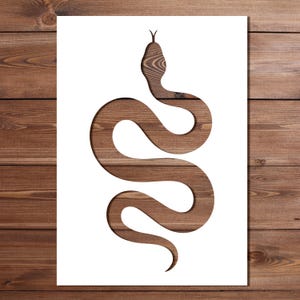 May include: A white rectangular stencil featuring a cutout in the shape of a winding snake. The snake silhouette is positioned vertically in the centre, revealing a dark brown wood grain pattern through the negative space of the cutout design.