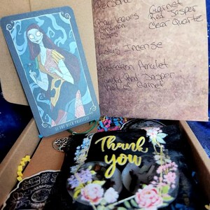 Puede incluir: Una pequeña caja llena de varios artículos, incluyendo una carta de tarot con un personaje de dibujos animados, una pequeña bolsa con un diseño floral y una nota escrita a mano que enumera los ingredientes para una poción de poder personal. La nota incluye el texto "Personal Power" y enumera ingredientes como "bay leaves", "cinnamon cloves", "garnet", "red jasper", "clear quartz", "lotus incense", "protection amulet", "tumbled red jasper", and "vial of garnet". La bolsa tiene el texto "Thank you" impreso en ella.