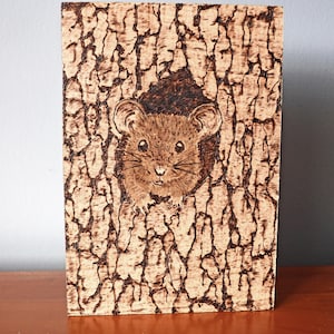 May include: A wooden plaque with a pyrography design. The artwork depicts a brown mouse emerging from a dark brown opening in a textured, bark-like background. The mouse has large ears and whiskers. The plaque is rectangular.