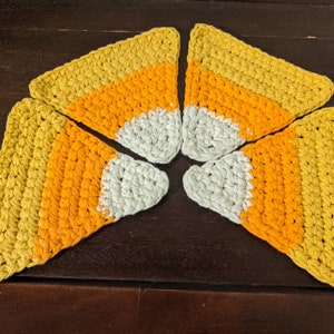 May include: Five crocheted candy corn shapes in yellow, orange, and white. The shapes are arranged in a circle on a brown wooden surface.