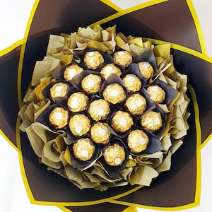 May include: A bouquet arrangement featuring numerous spherical chocolates wrapped in shiny gold foil. The chocolates are nestled within layers of dark brown and translucent gold tissue paper, framed by a dark brown outer wrap with yellow edges.
