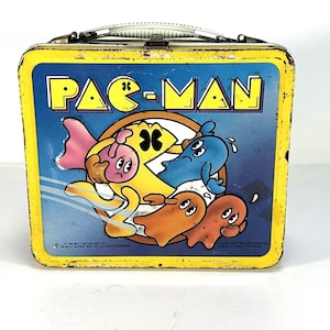 May include: Vintage Pac-Man lunchbox with a yellow border and a blue background. The lunchbox features the Pac-Man logo and colorful characters. The lunchbox has a metal handle and a clasp closure. The lunchbox is a retro collectible.