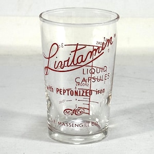 May include: A clear glass measuring cup with red lettering that reads "Livitamin Liquid Capsules with Peptonized Iron" and "The Massengill Co."