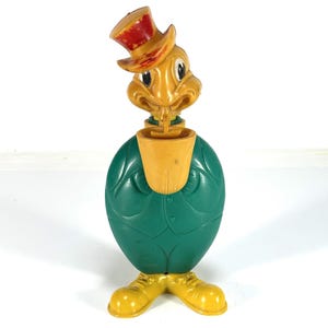 May include: A vintage plastic toy figure of a cartoon character. The figure has a yellow head with a red-topped hat, a green body, and yellow shoes. The figure is standing upright and appears to be a collectible item.