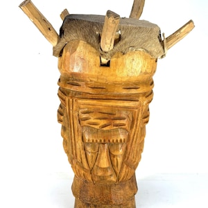 May include: A carved wooden sculpture with a cylindrical shape, featuring intricate facial carvings and a textured, fur-covered top. Four wooden rods extend from the top. The sculpture is made of light brown wood.