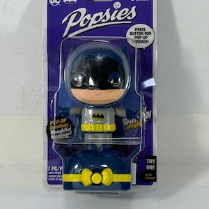 May include: A blue and yellow Batman Popsies figure with a pop-up message feature. Press the button on the base to reveal a hidden message. The figure is packaged in a clear plastic blister pack with a purple background and the Funko logo.