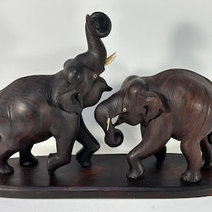May include: Two dark brown wooden elephants facing each other, one with its trunk raised, on a dark brown wooden base.