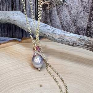 May include: A gold-colored chain necklace with a large, pearlescent, lavender-colored pearl pendant. The pendant is accented with small pink gemstones. The necklace is displayed on a light-colored wooden surface.