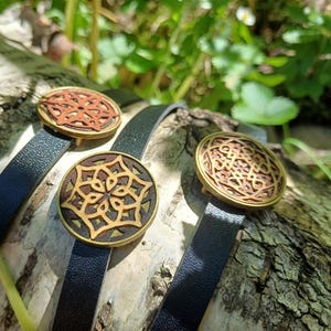 Könnte beinhalten: Drei Lederarmbänder mit runden Holzanhängern mit komplizierten geometrischen Mustern. Die Anhänger sind in Braun- und Rottönen gehalten, die Armbänder sind schwarz.