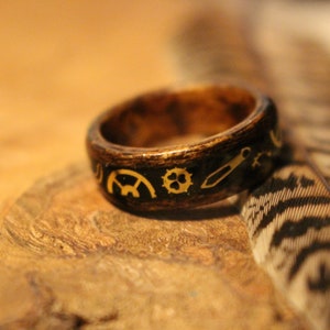 Può includere: Un anello in legno marrone scuro con dettagli dorati. L'anello è decorato con piccoli disegni intricati, tra cui ingranaggi e altri elementi meccanici.