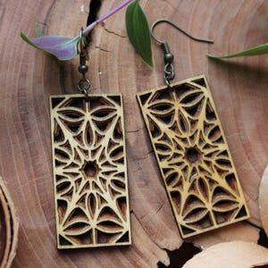 Peut inclure: Paire de boucles d'oreilles en bois avec un motif géométrique. Les boucles d'oreilles rectangulaires présentent un motif ajouré en couches avec une étoile centrale et des formes florales. Elles sont suspendues à des crochets d'oreilles de couleur bronze.