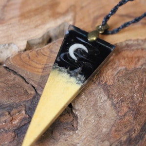 Op de afbeelding: Een zwart-witte hars hanger met een halve maan ontwerp, hangend aan een zwart koord. De hanger is driehoekig en heeft een lichte, hout-achtige basis.